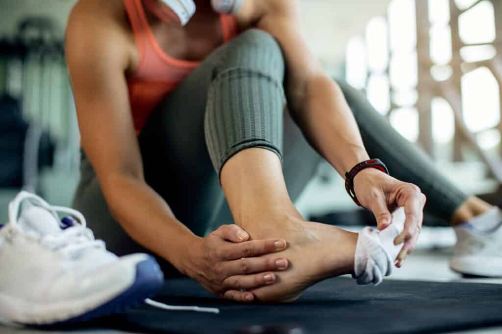 Close up image of a person in workout attire sitting on the gym floor, holding their right ankle and reflecting on the anticipated sprained ankle healing time.