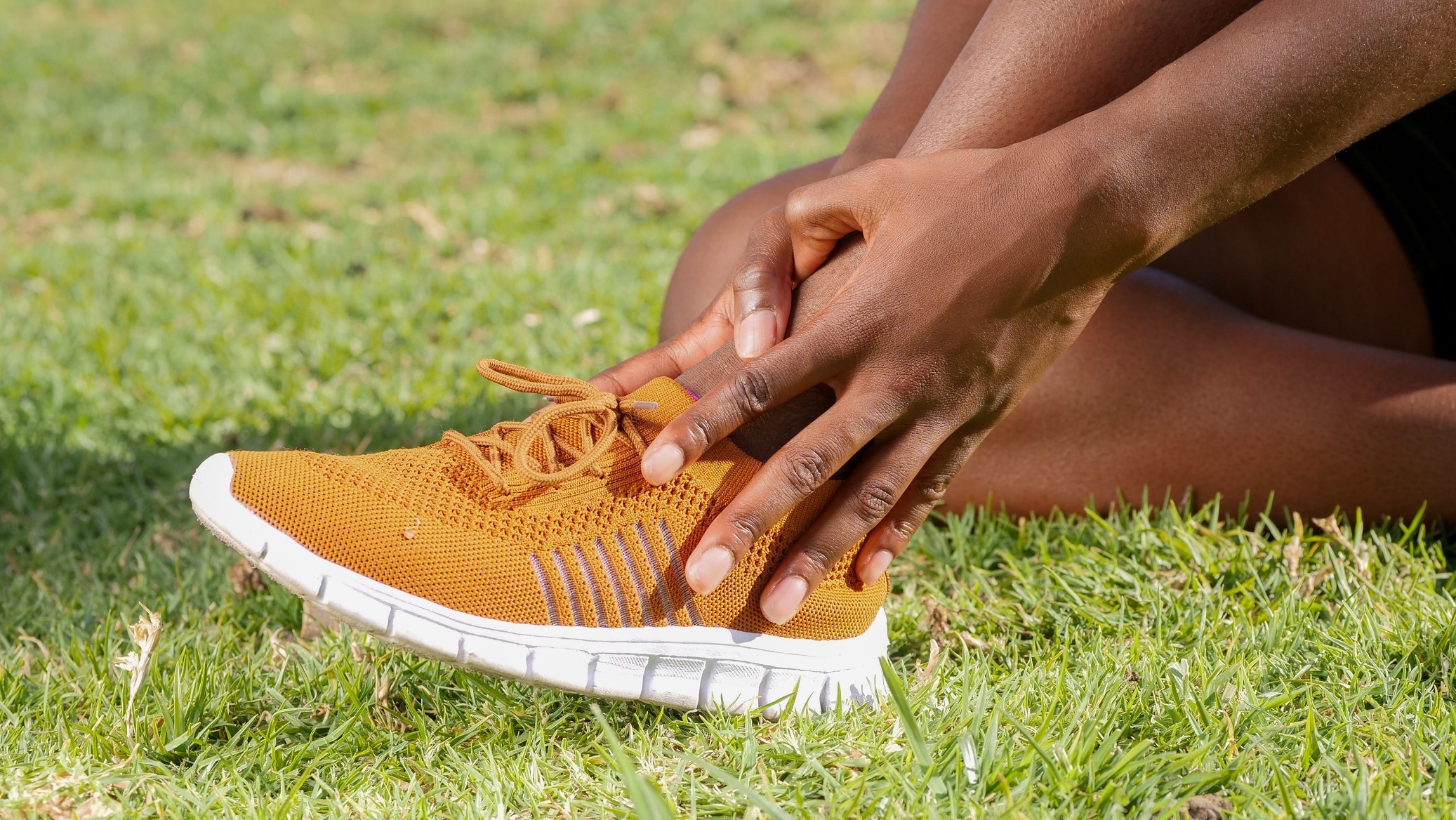 Close up image of a person sitting in the grass, wearing dark yellow running sneakers and grasping their left ankle, contemplating sprained ankle heal time.