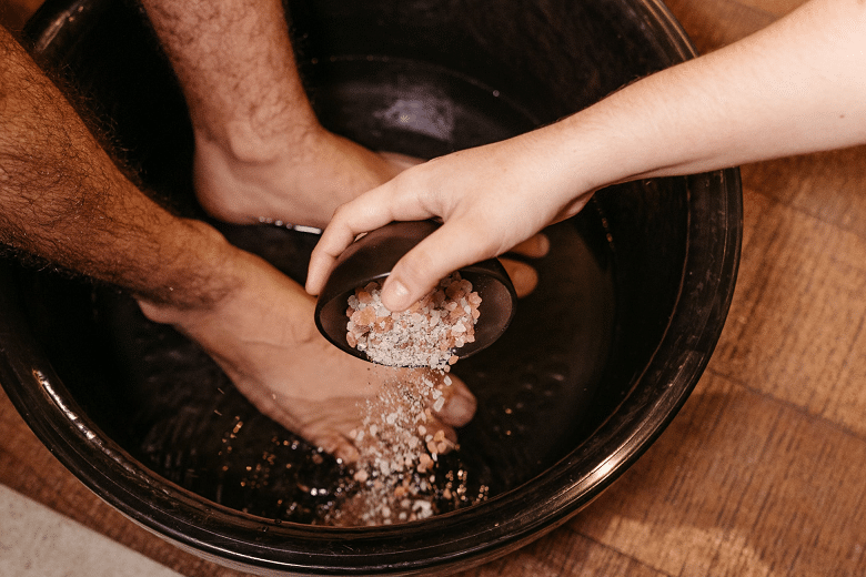 Close-up image of a person’s feet in a bucket of water as another person’s hand dumps epsom salt into the water, depicting pain management and its role in sprained ankle healing time.