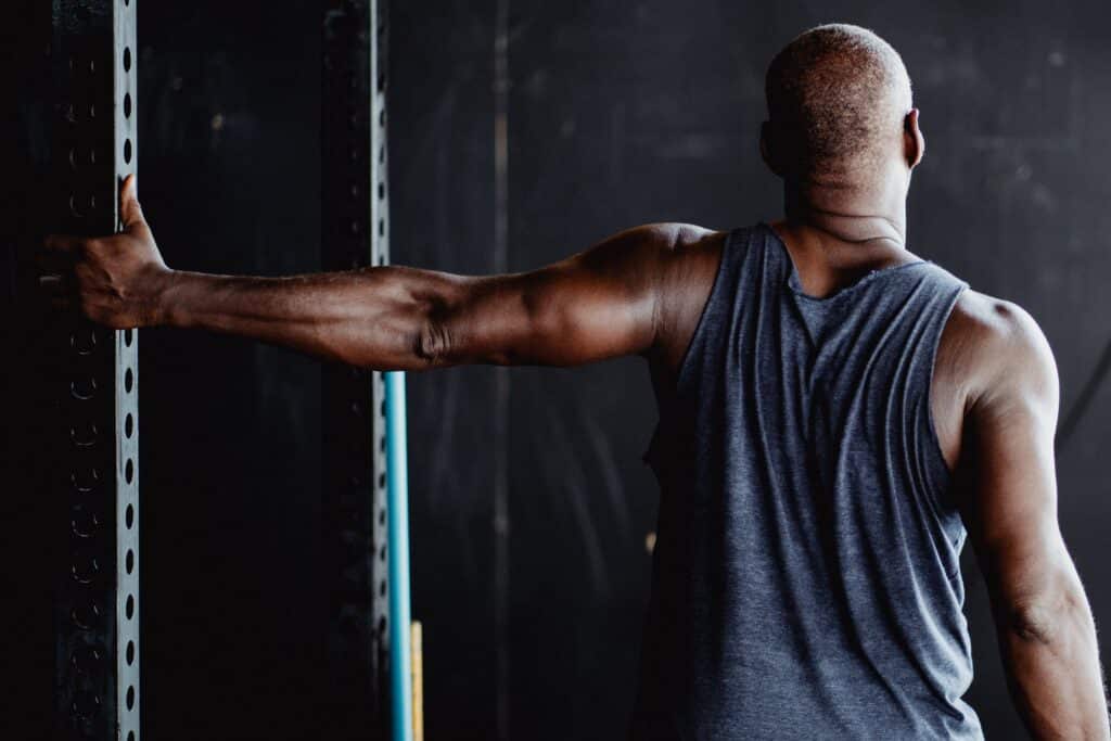 A person is standing in a gym with their right arm by their side and their left arm grasping a vertical metal beam, stretching their shoulder as if alleviating rotator cuff pain.