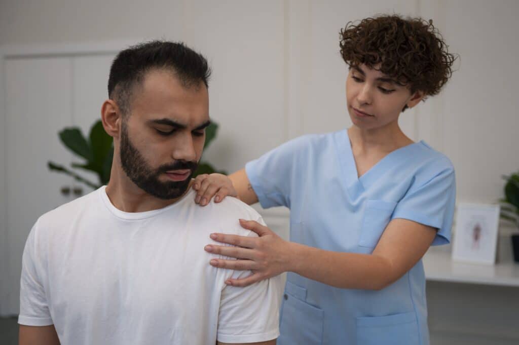 A person dressed in scrubs is examining a patient’s left shoulder with both hands; the patient is looking at their shoulder with a slightly pained expression, suggesting rotator cuff pain.