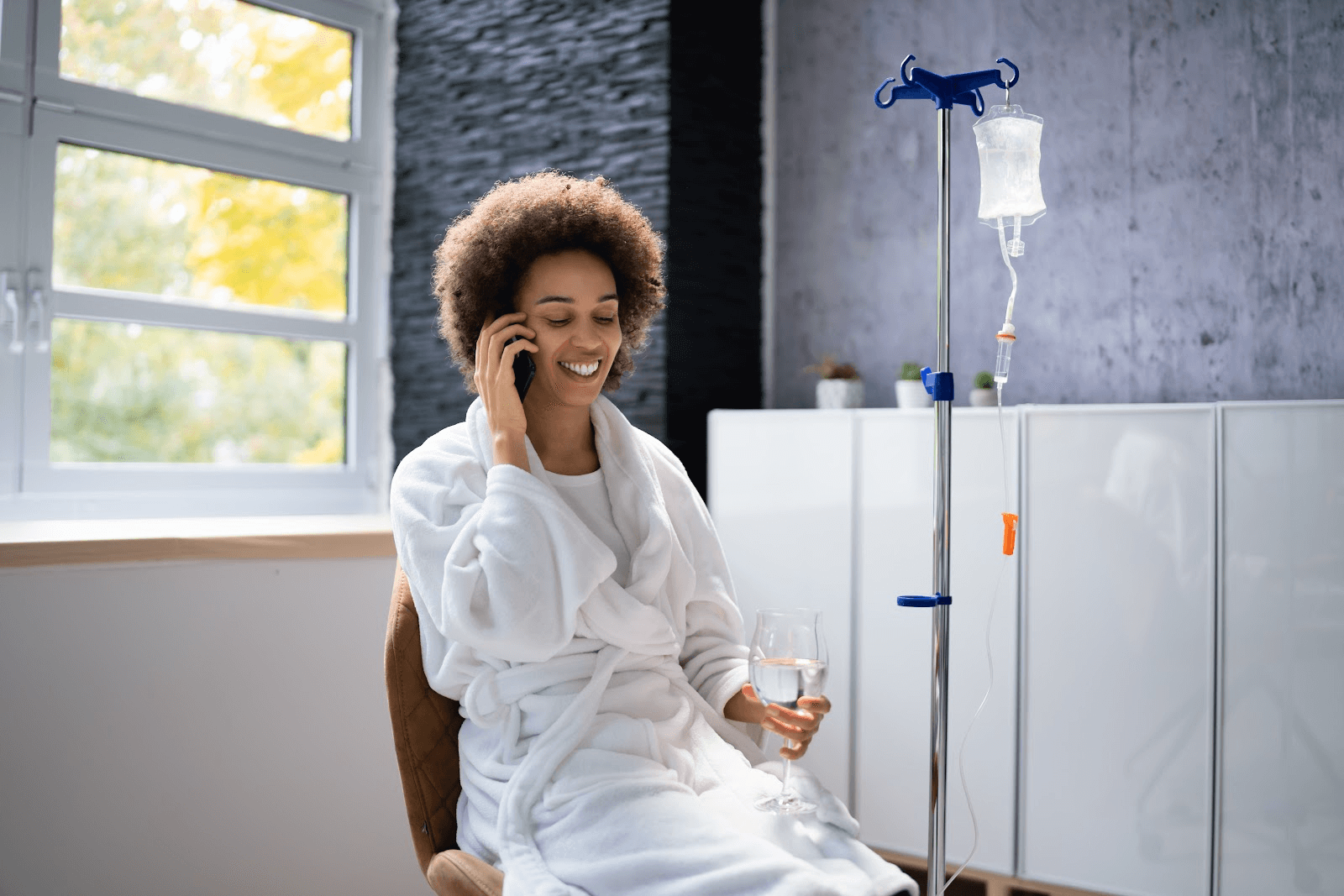 Vitamin C IV Therapy; image of a relaxed woman wearing a spa robe and talks on the phone while receiving an IV drip.