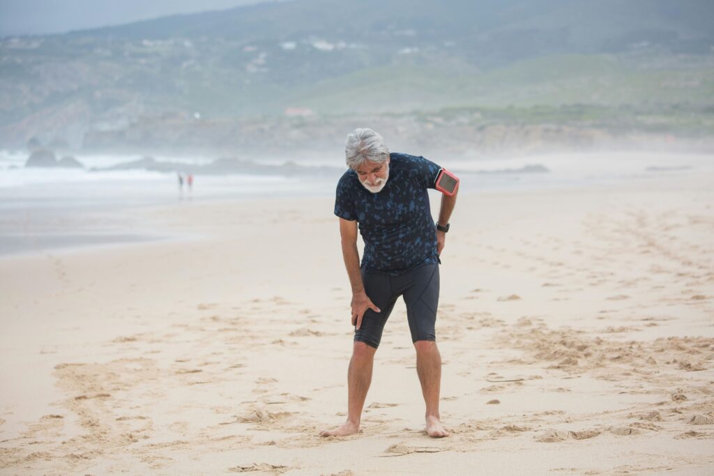 Photo of a person standing on a beach, slightly hunched over with one hand on their leg and one on their back, indicating sciatica pain.