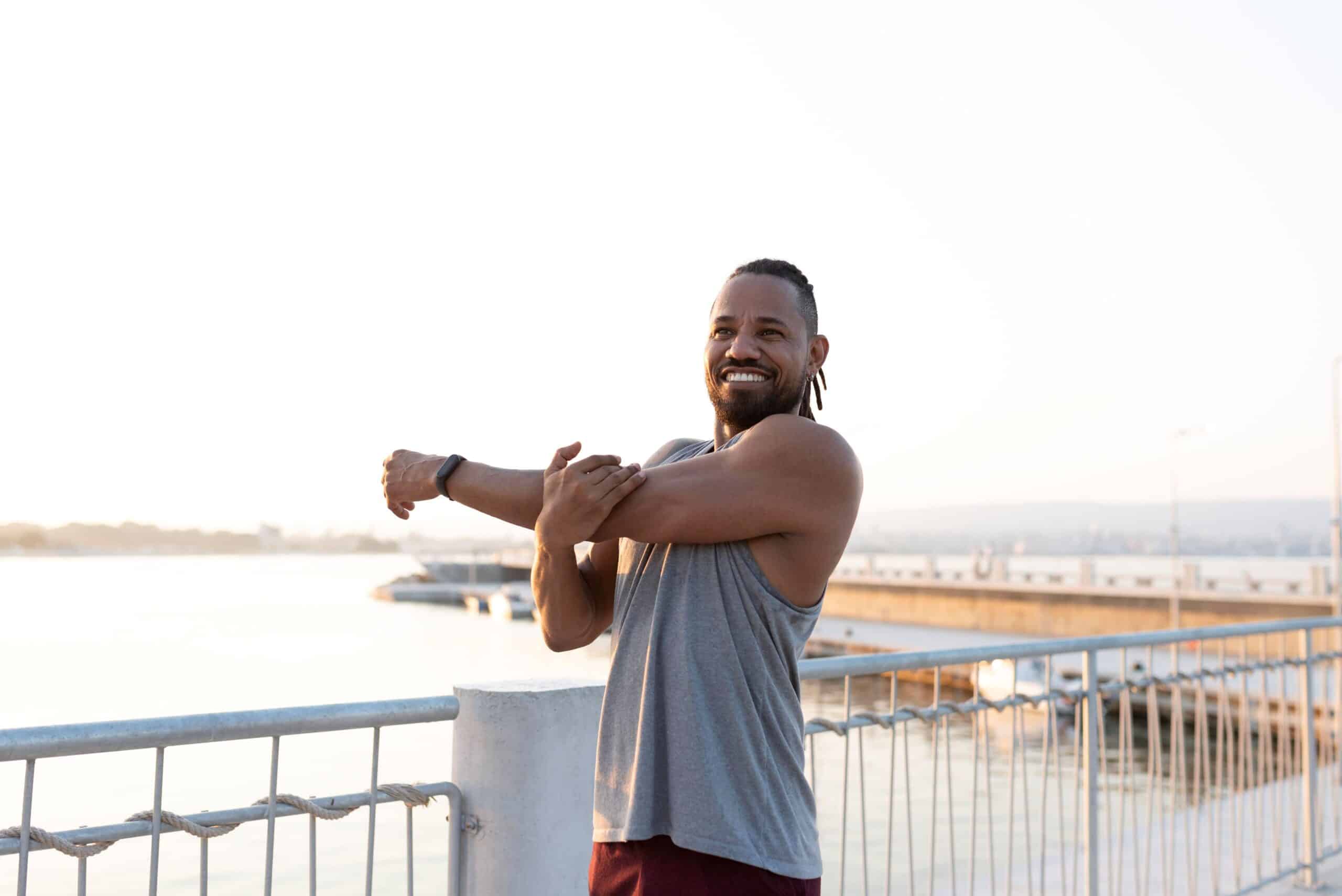 Frozen shoulder; image of a happy man wearing a gray tank top out on the marina stretching his left shoulder.