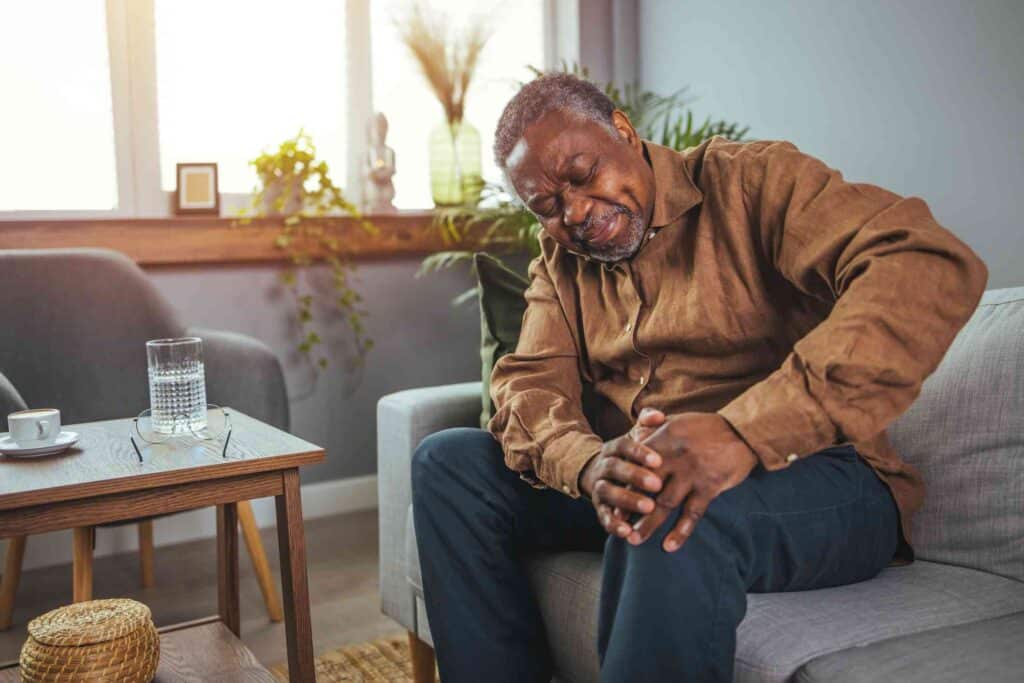 PRP injection for knee; image of an elderly man sitting on a gray couch and grasping his left knee with both hands in pain.