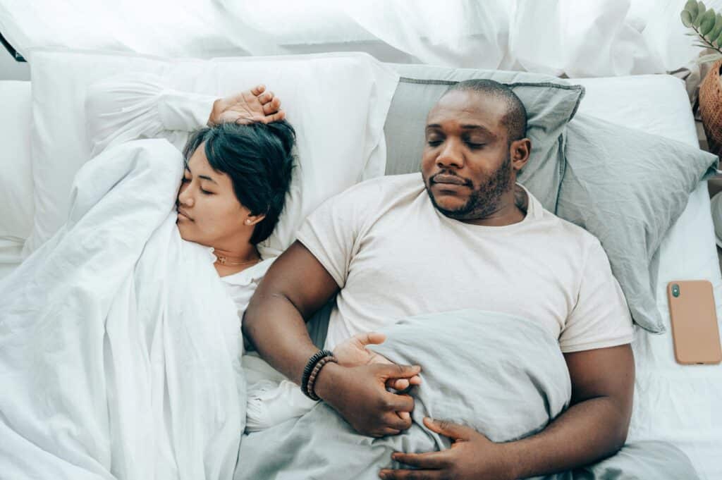 neck pain from sleeping, Image of a couple sleeping in bed together, on their backs, the woman sleeping on the left side of the bed and the man sleeping on the right.