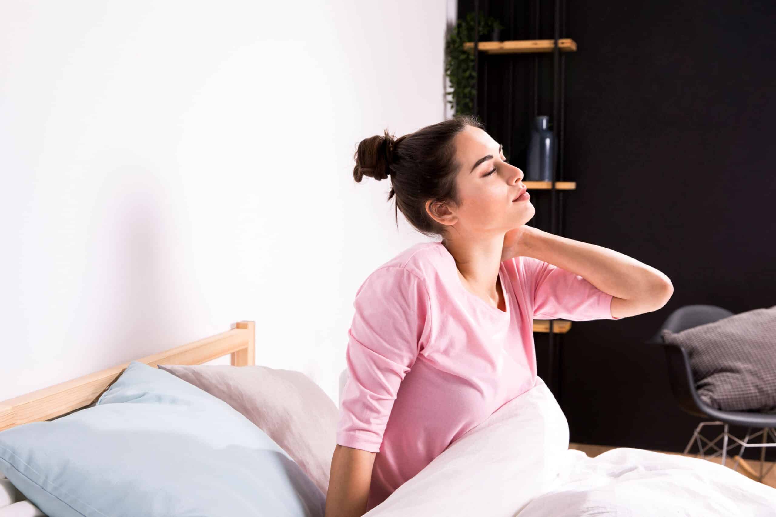 neck pain from sleeping, Image of a woman sitting up in bed with neck pain, grasping her neck with her left hand.