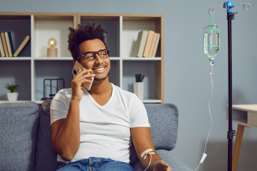 Image of a happy man sitting on the couch and talking on the phone while receiving IV wellness treatment.