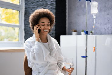 Image of a happy female patient receiving an IV Wellness Therapy treatment while talking on the phone and holding a glass of water.