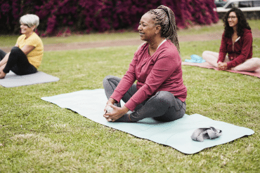 Image of three woman participating in an outdoor yoga class, sitting in a yoga pose on their yoga mats.