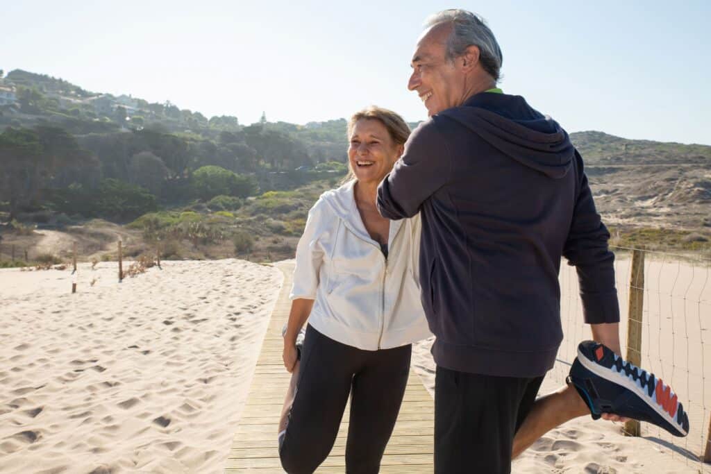 Orthopedic stem cell therapy, image of an older man and woman couple stretching their legs and knees on a beach boardwalk.