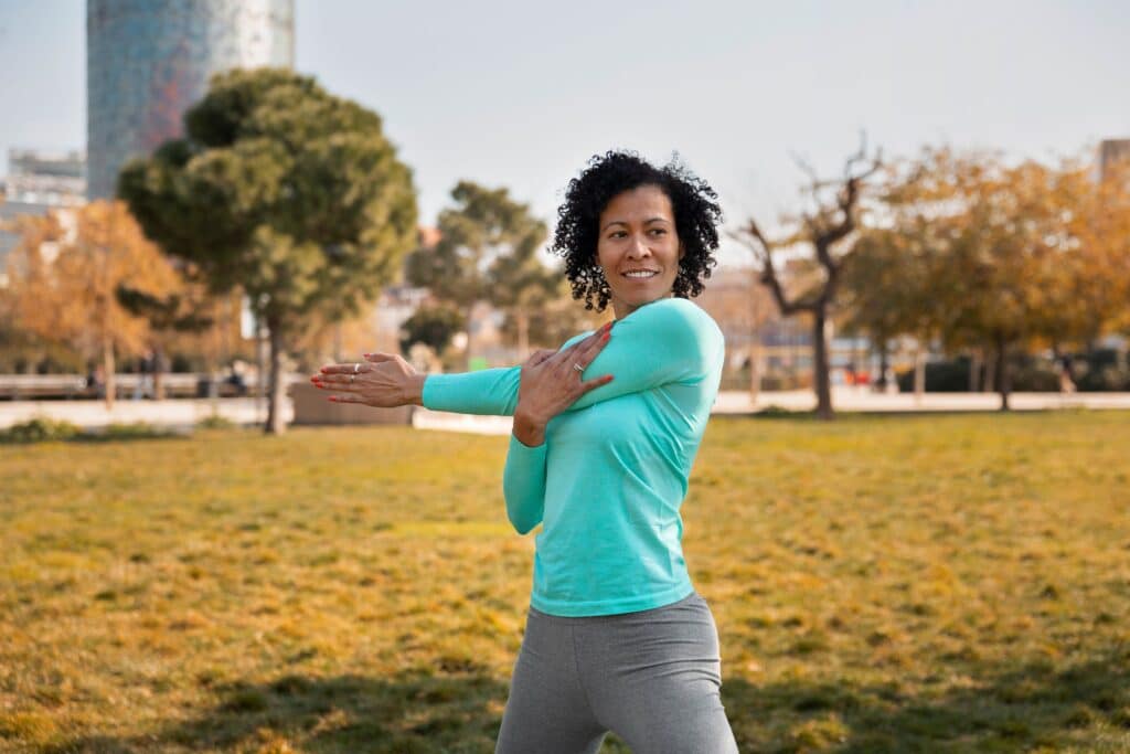 Unnecessary spine surgeries, image of a woman standing and stretching their left shoulder outside at the park.