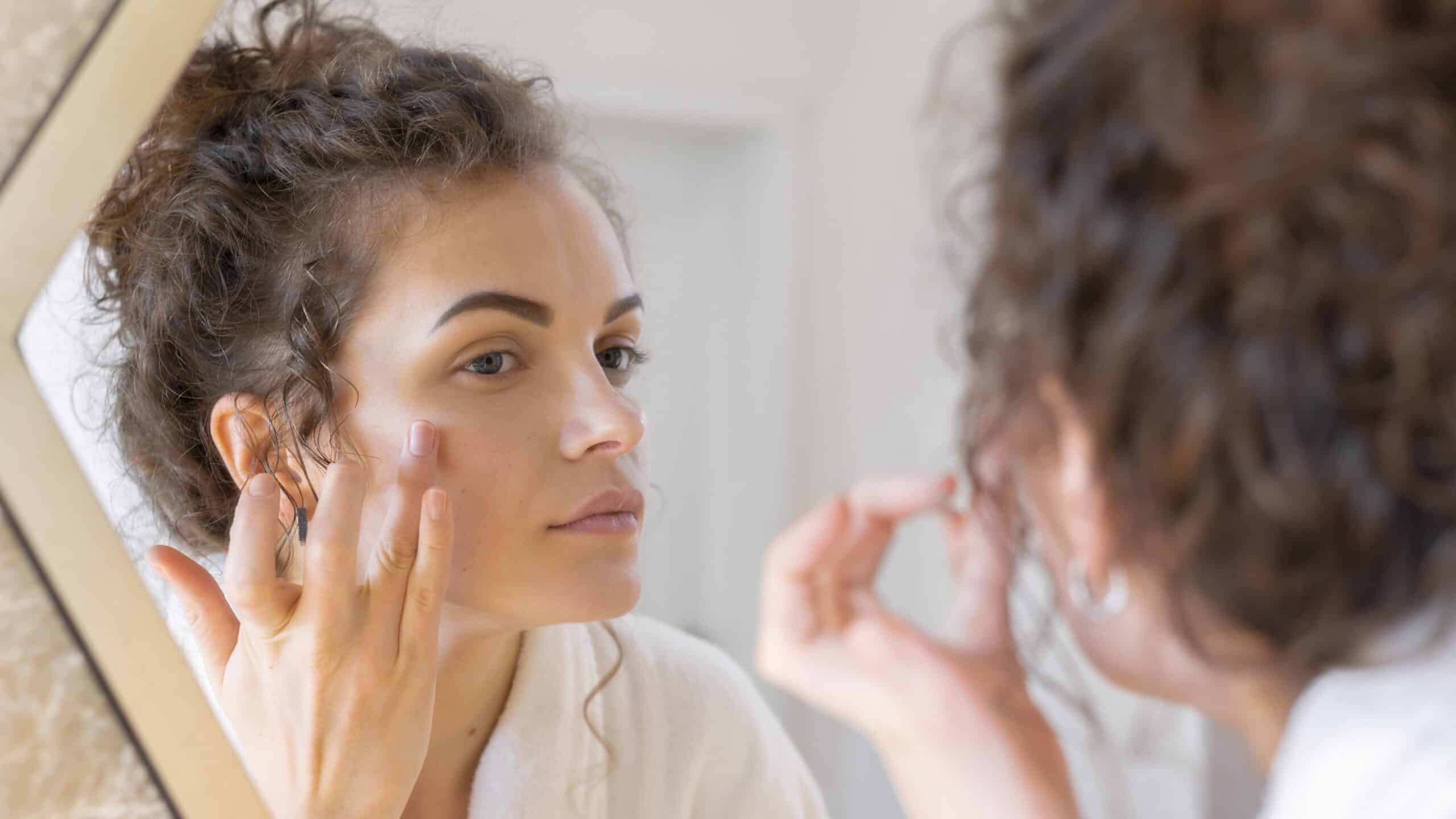 What is a Vampire Facial®, image of a young woman examining her face in the mirror and touching her face with her left hand.