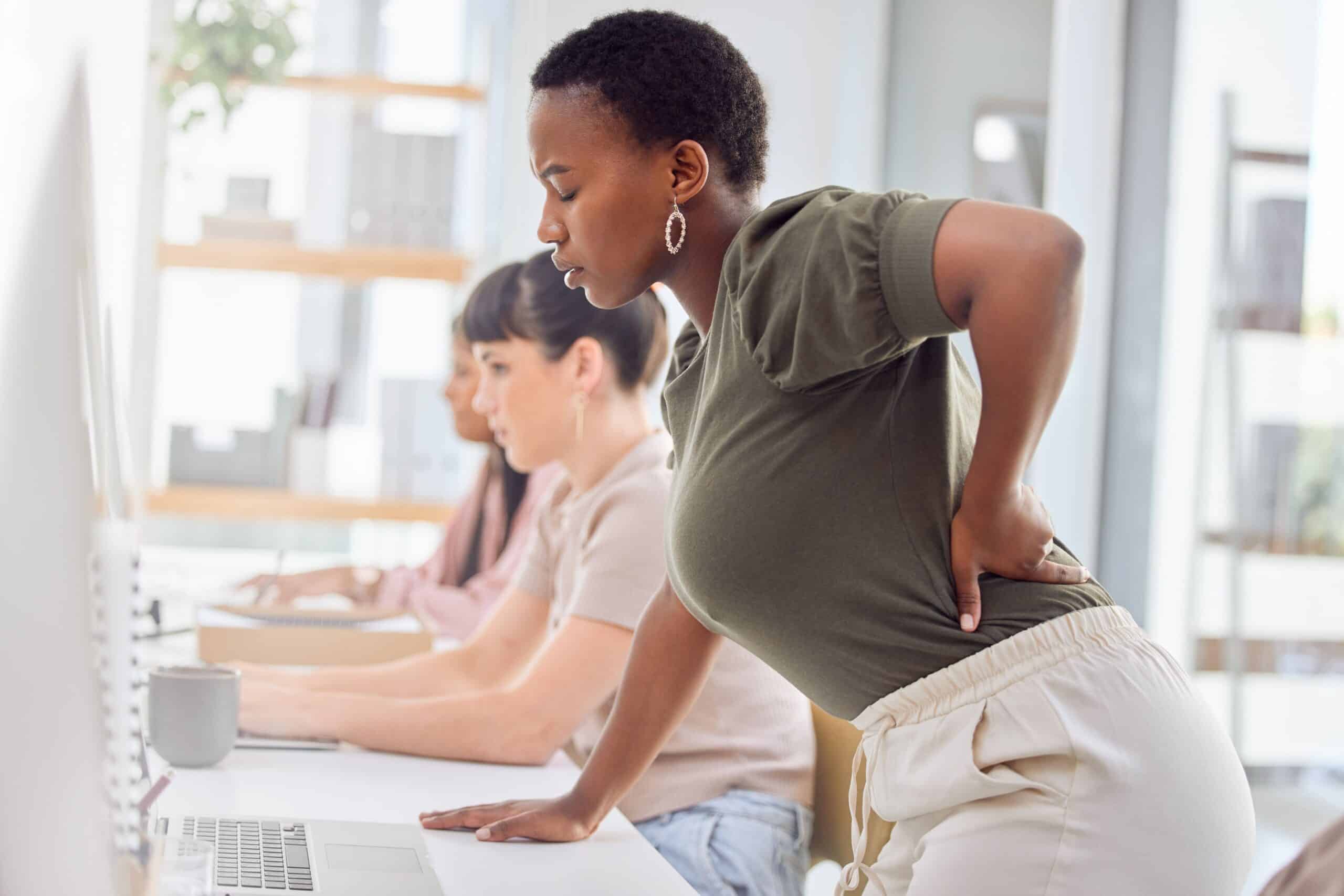 Can Adhesions Cause Back Pain, image of a woman wearing an olive green blouse and white pants standing at her desk, leaning forward and grasping her lower back with her left hand.