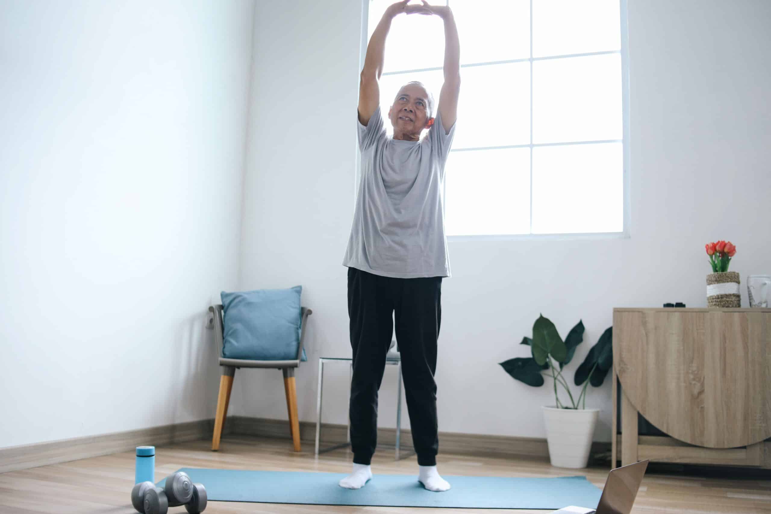 Can Adhesions Cause Back Pain, image of an elderly man wearing a gray t-shirt and black pants stands on a blue yoga mat and stretches both arms over his head.