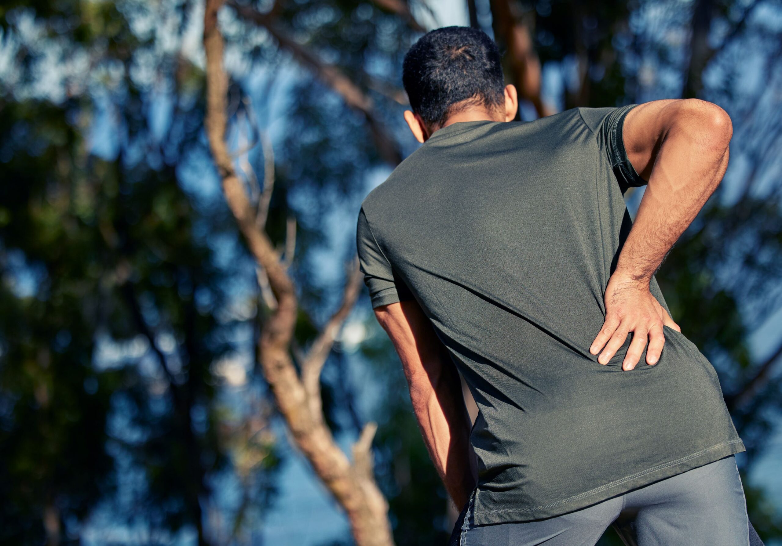 Can Adhesions Cause Back Pain, image of the back view of a man with dark hair, wearing an olive green t-shirt, bending forward and grasping his lower back with his right hand.