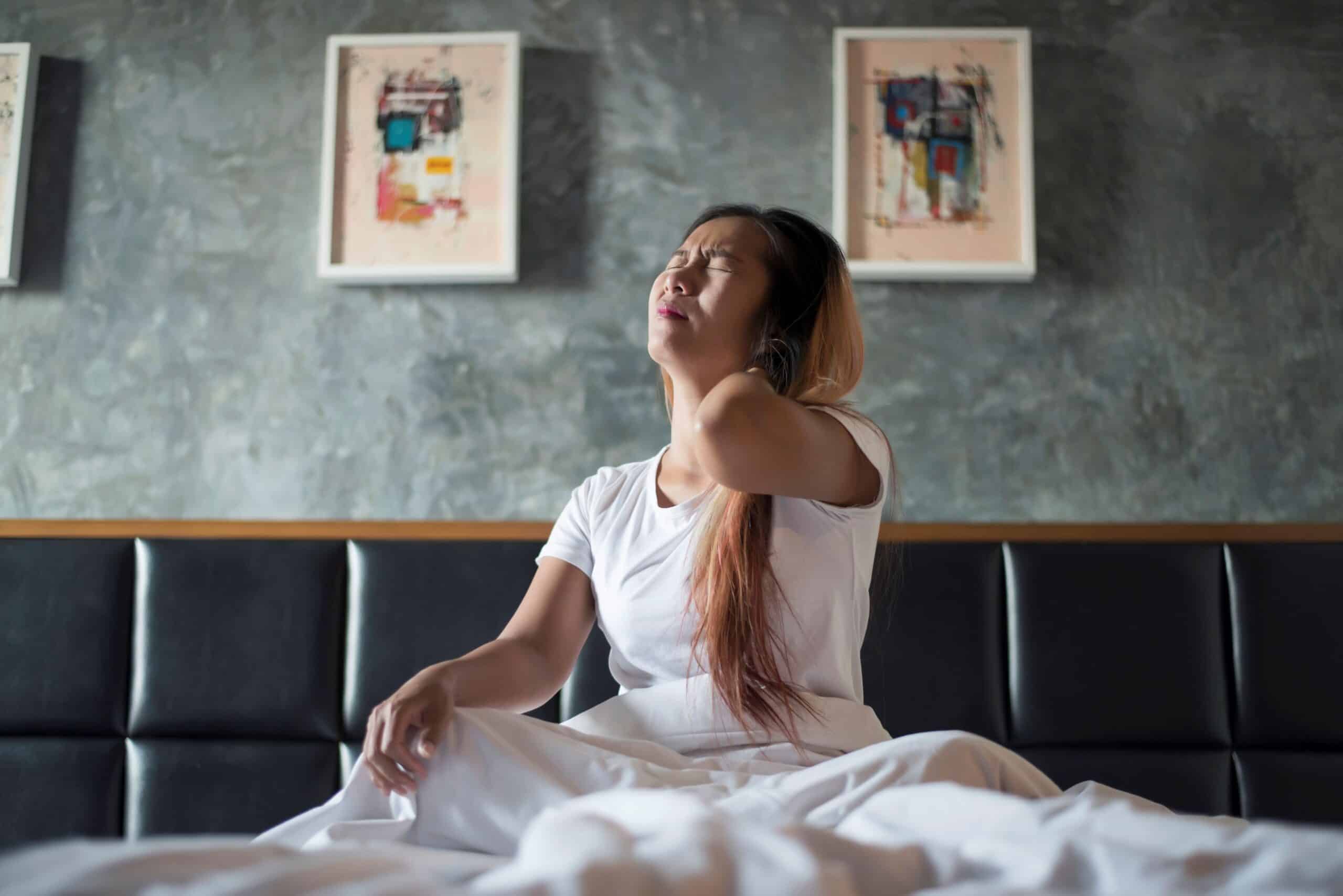 How To Sleep With Cervical Radiculopathy, image of a woman sitting up in bed with a look of pain on her face, grasping the back of her neck with her left hand.