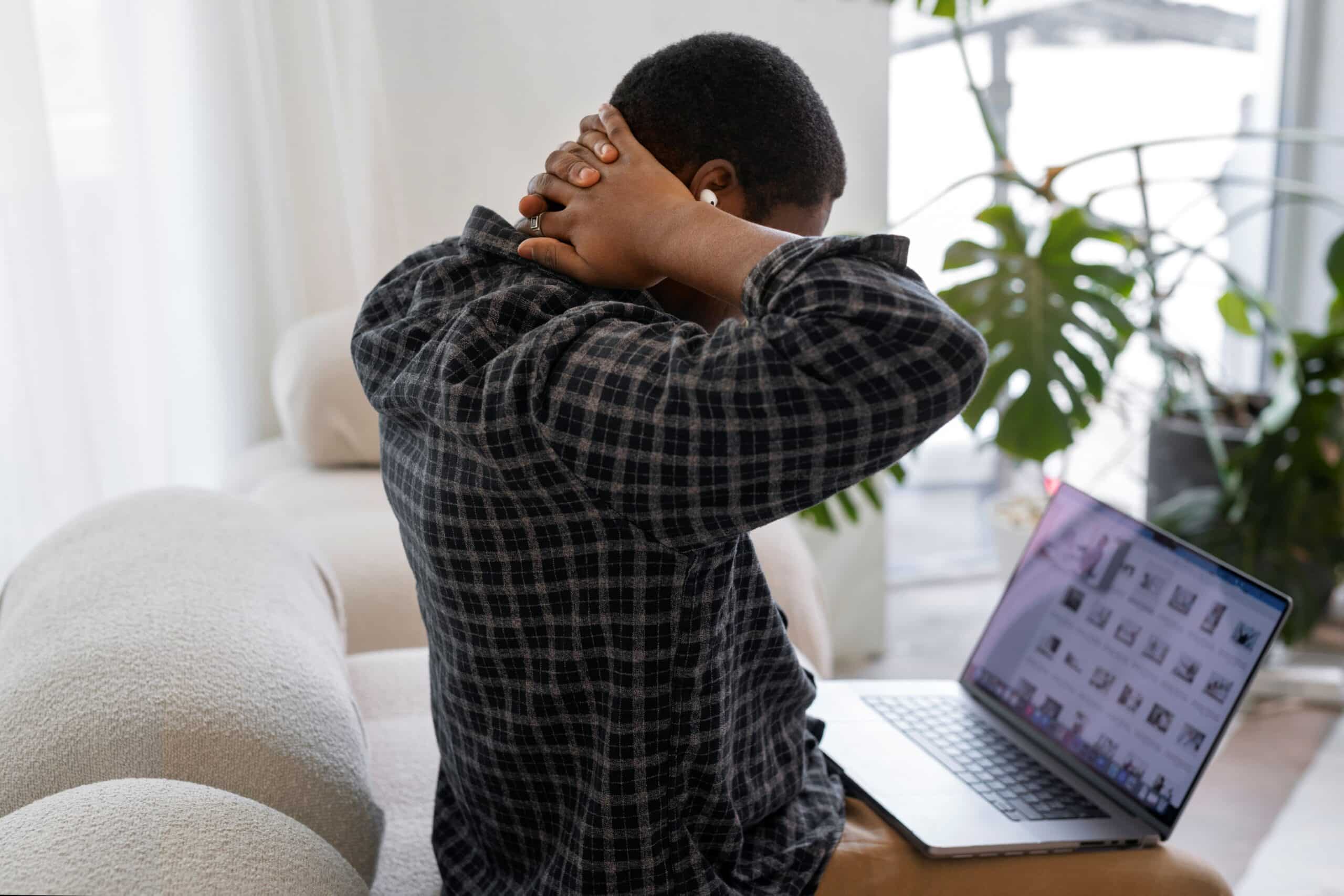 How To Sleep With Cervical Radiculopathy, image of a man sitting on a couch with a laptop in his lap looking down with both his hands clasped behind his head, on his neck.