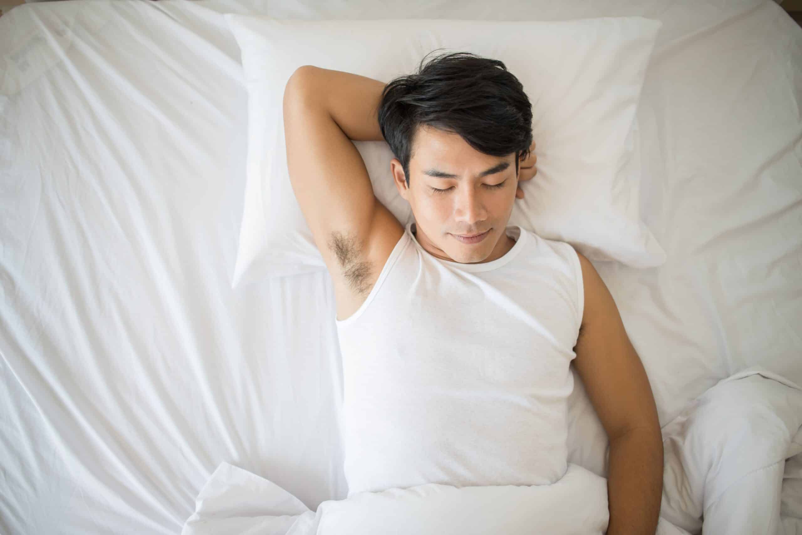 How To Sleep With Cervical Radiculopathy, image of a man in a white tank top on a white bed sleeping on his back with his right arm tucked behind his head.
