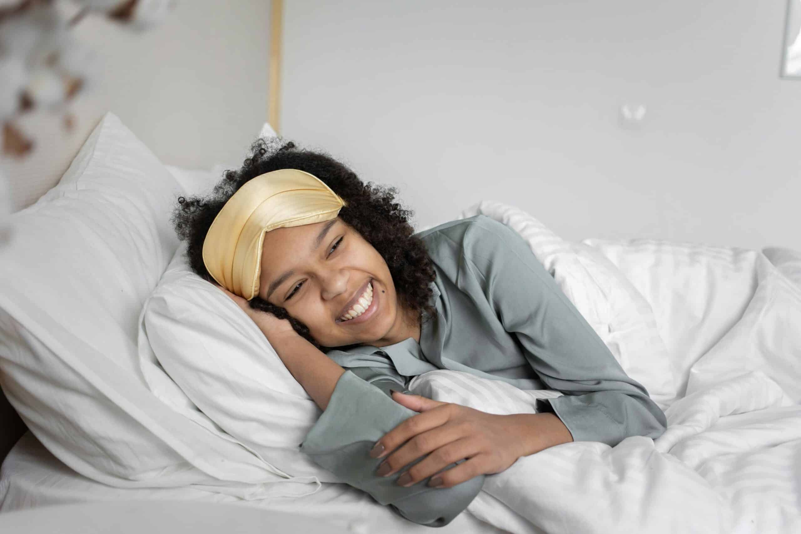 How To Sleep With Cervical Radiculopathy, image of a happy woman lying in bed on her right side with a sleep mask worn across her forehead.