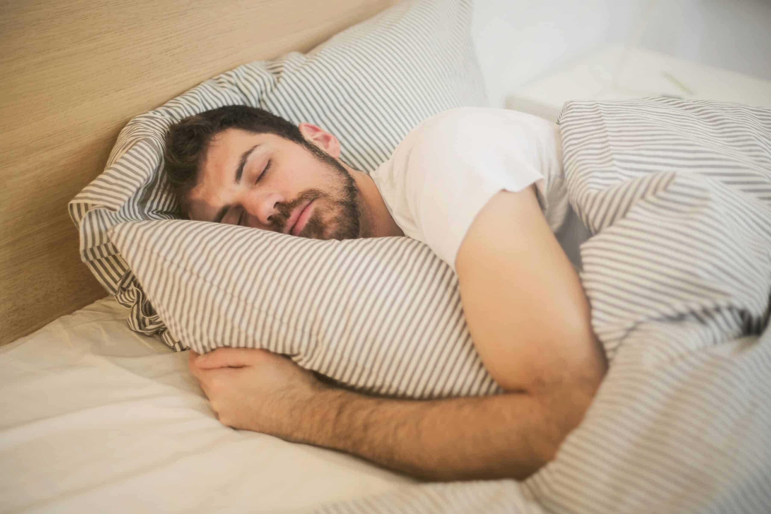How To Sleep With Cervical Radiculopathy, image of a man sleeping in bed on his left side, embracing a pillow with a white and gray stripe cover.