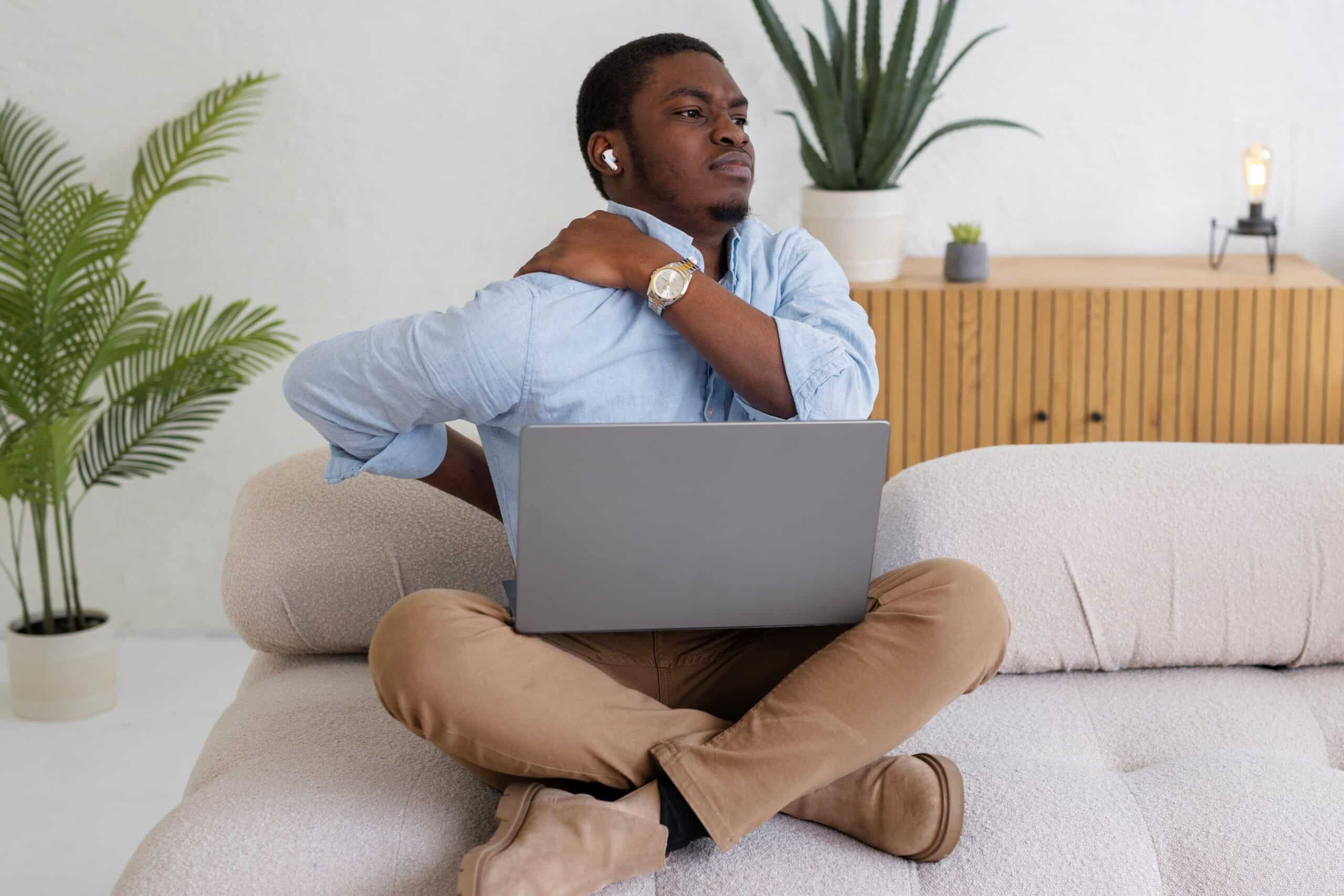 Back pain from sitting at desk, image of a man sitting on a couch with his legs crossed and a laptop in his lap, grasps his lower back with his right hand and grasps his right shoulder with his left hand.