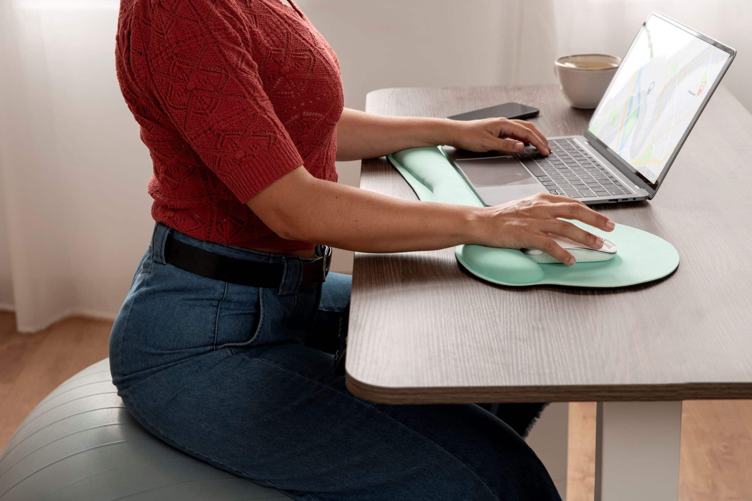Back pain from sitting at desk, image of a woman with good posture sitting on a big inflatable ball at her desk, using her laptop and mouse.