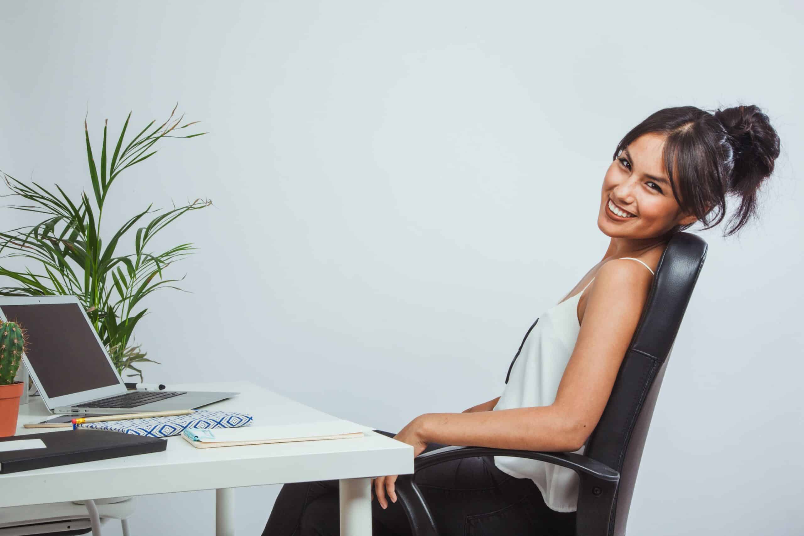 Back pain from sitting at desk, image of a smiling woman sitting back in her office chair at her desk with her laptop open.