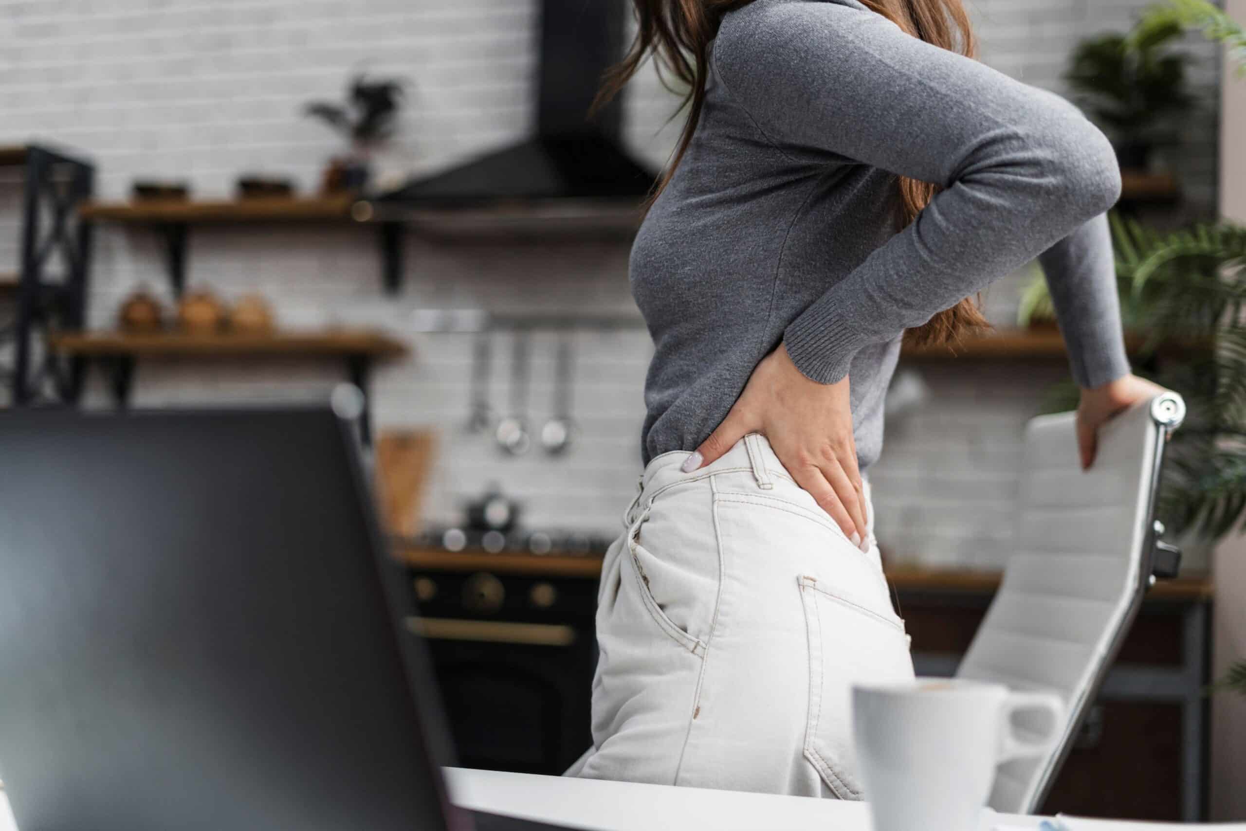 Back pain from sitting at desk, image of a woman standing up at her desk, leaning on her white office chair with her left hand grasping her lower back.
