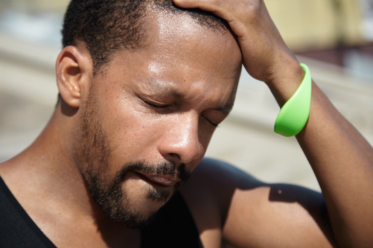 Can Heat Cause Headaches, image of a man with a look of pain on his face and a green bracelet around his left wrist has left hand placed on his head.