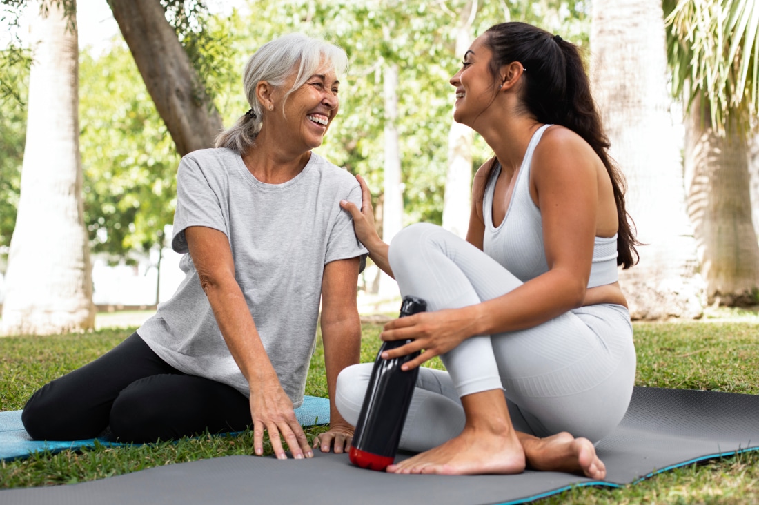 Can Heat Cause Headaches, image of a happy young woman and happy older woman sitting on yoga mats in the park, smiling at each other.
