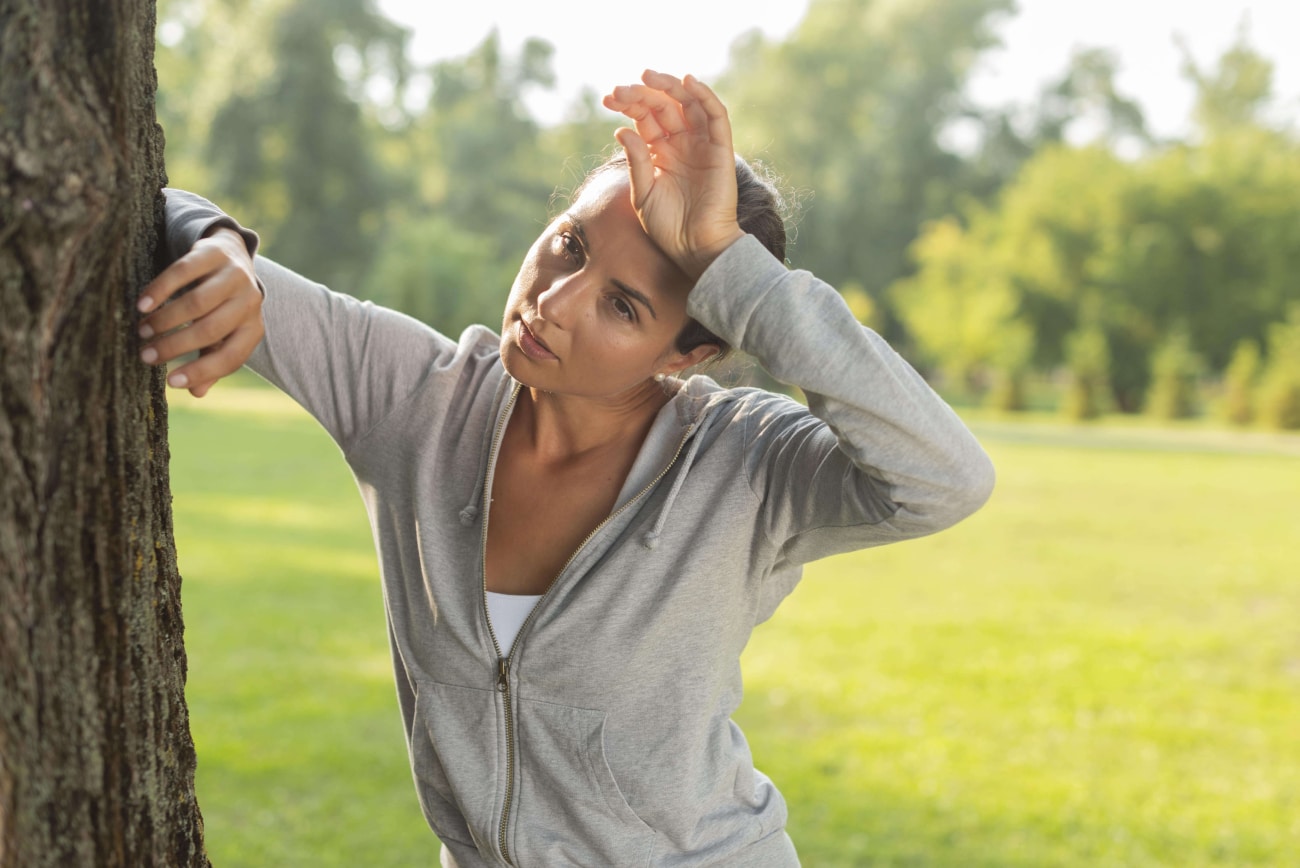 Can Heat Cause Headaches, image of a tired woman wearing a gray zip-up hoodie, leaning on a tree with her left hand resting on her forehead.