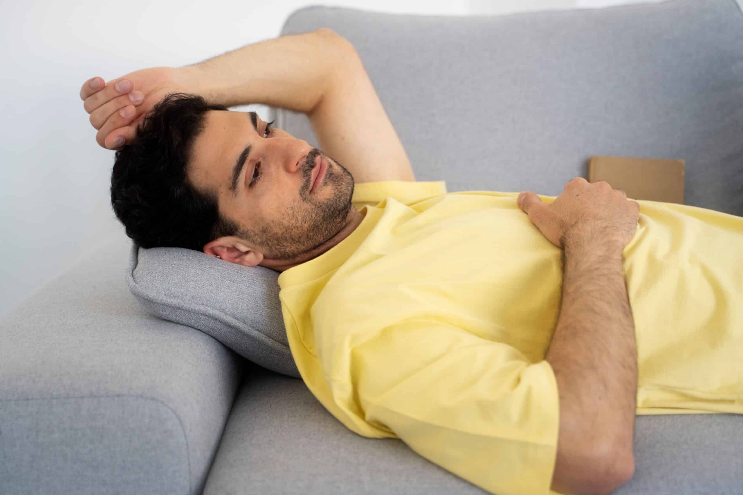 Why does my back hurt when I lie down, image of a man lying on his back on a gray couch with his right hand resting on his chest and his left hand resting on his head.