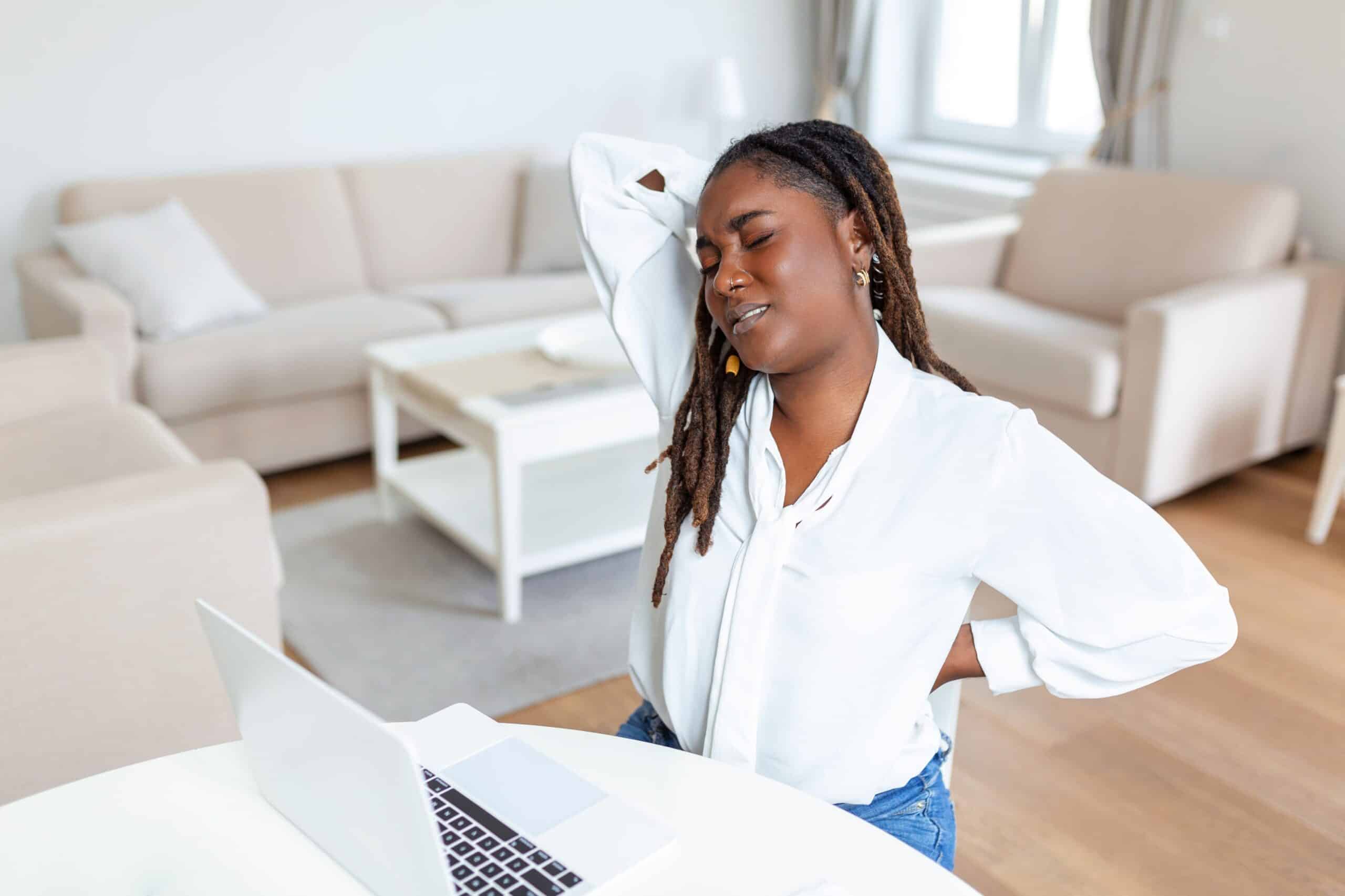 Why does my back hurt when I lie down, image of a woman sitting at a white table with her lap top in front of her grasps her back with both hands with a look of pain across her face.