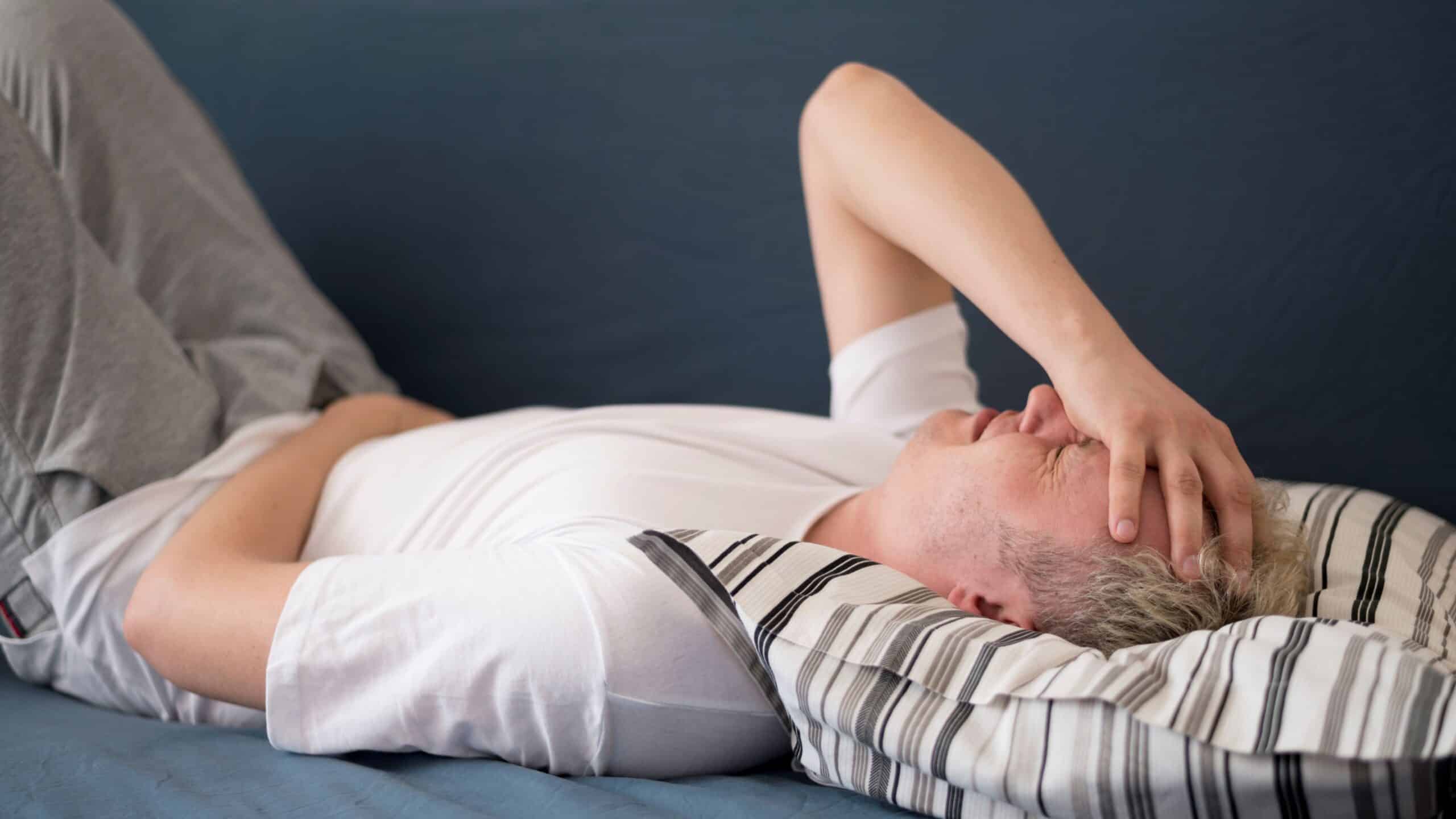 Why does my back hurt when I lie down, image of a man lying on his back with his knees bent, his right hand over his face, and a look of pain across his face.