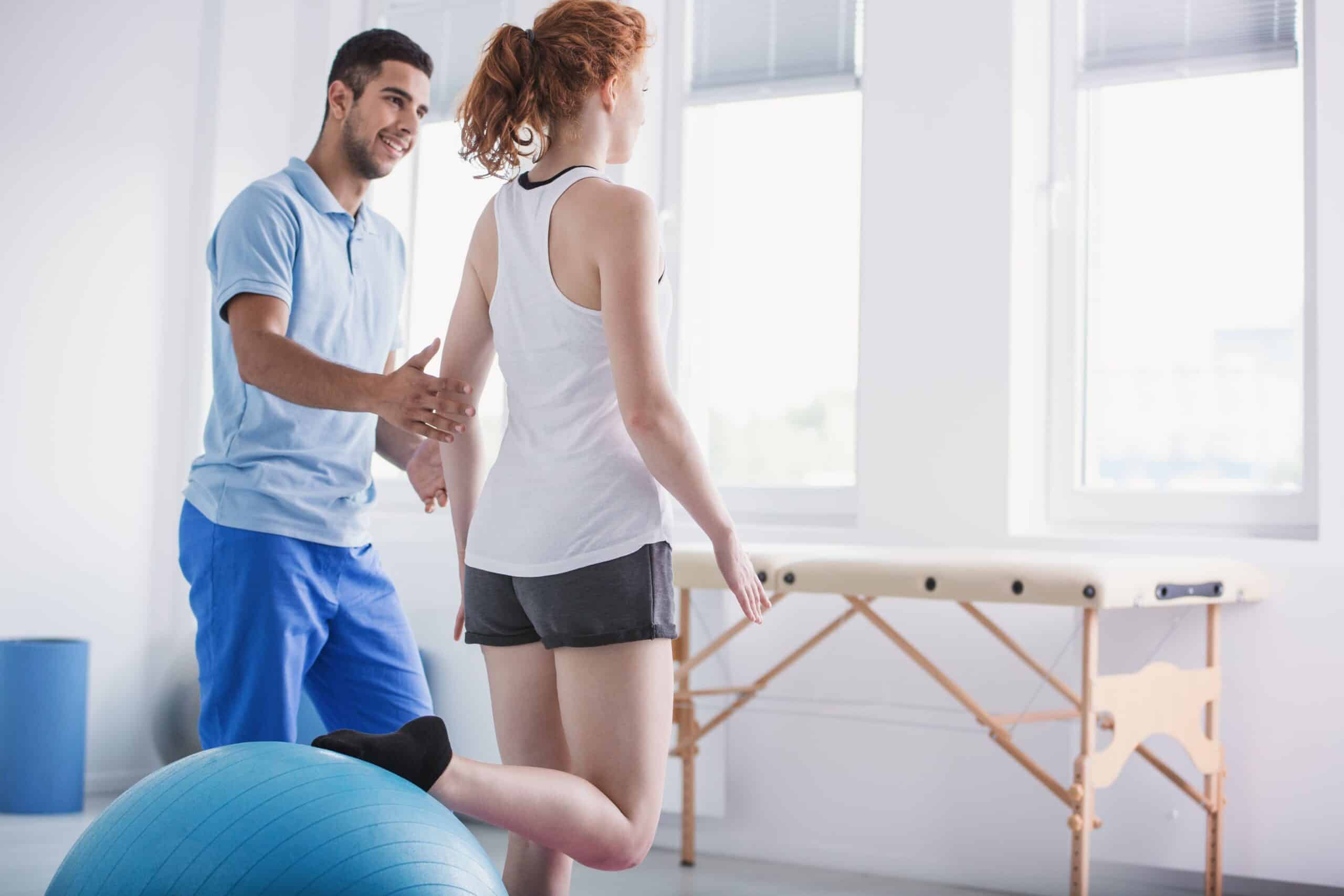 Gait training, image of a young male physical therapist helping a female patient steady herself as she stands and balances with her right knee bent and foot placed behind her on a big blue medicine ball.
