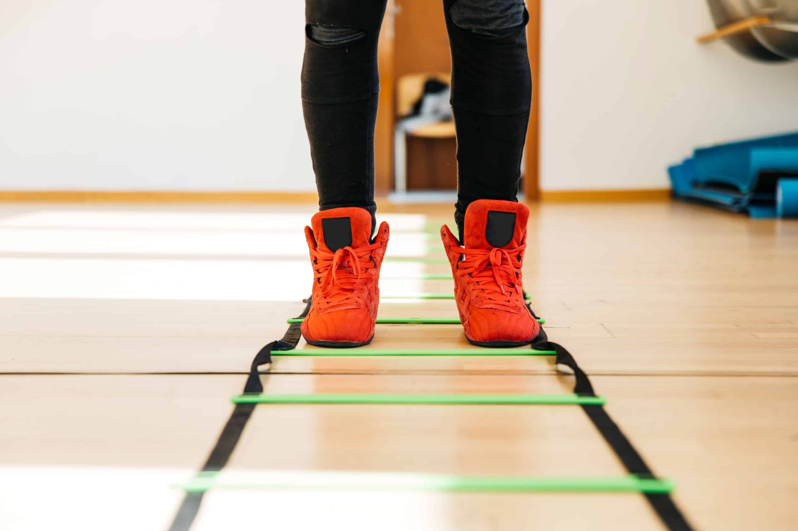 Gait training, image of a person wearing bright orange sneakers and black workout pants stands in the middle of obstacle lines placed on a gym floor.