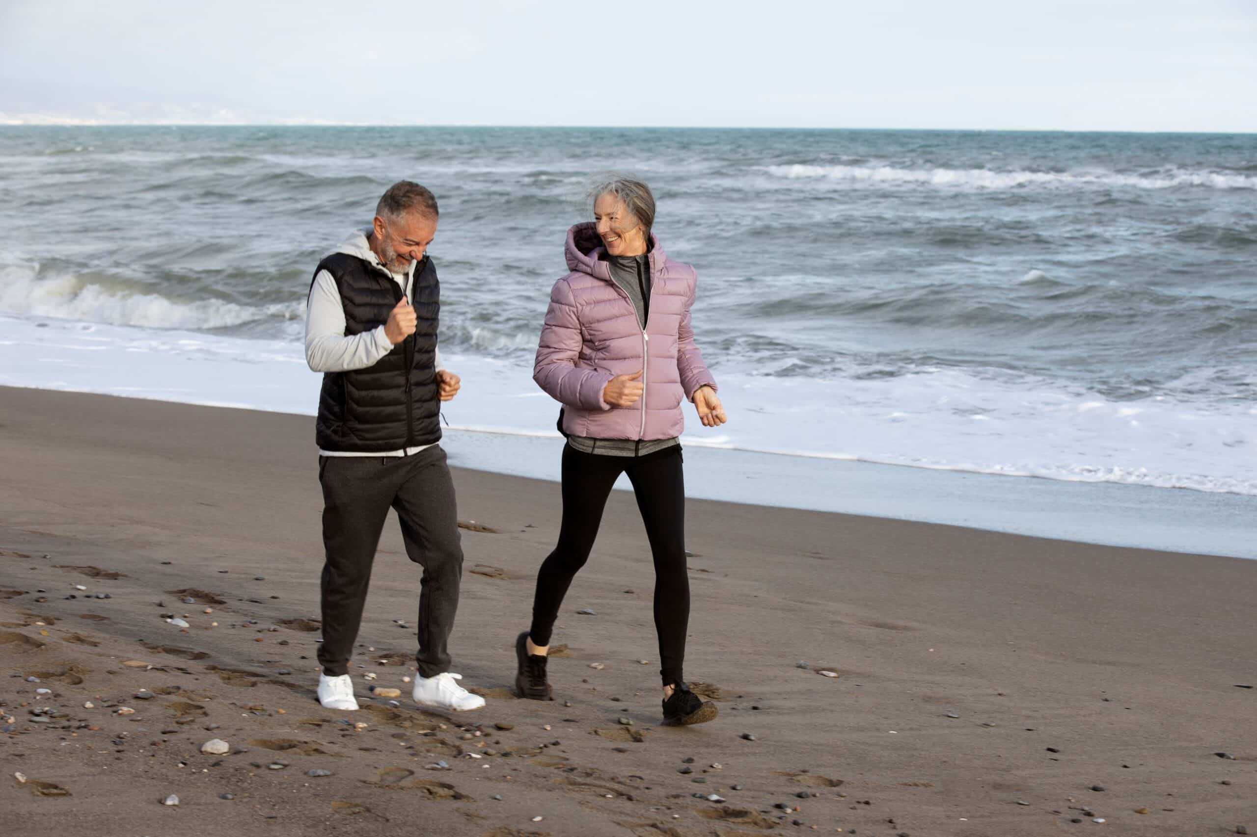 Gait training, image of a smiling older couple wearing warm workout clothes walking at a fast pace along a beach.