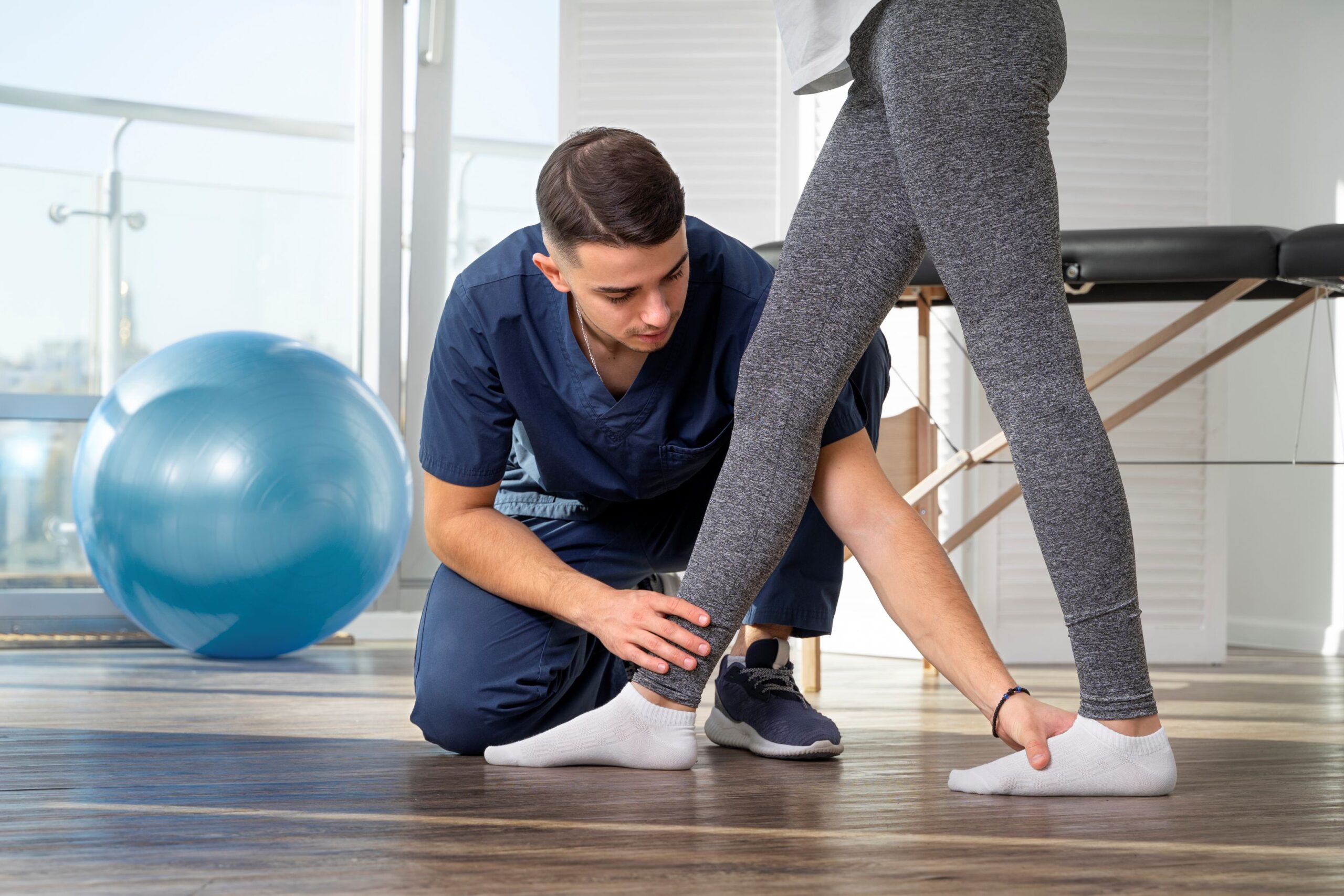 Gait training, image of a young male physical therapist wearing dark blue scrubs places one hand on a female patient’s right ankle and the other hand on the patient’s left foot.
