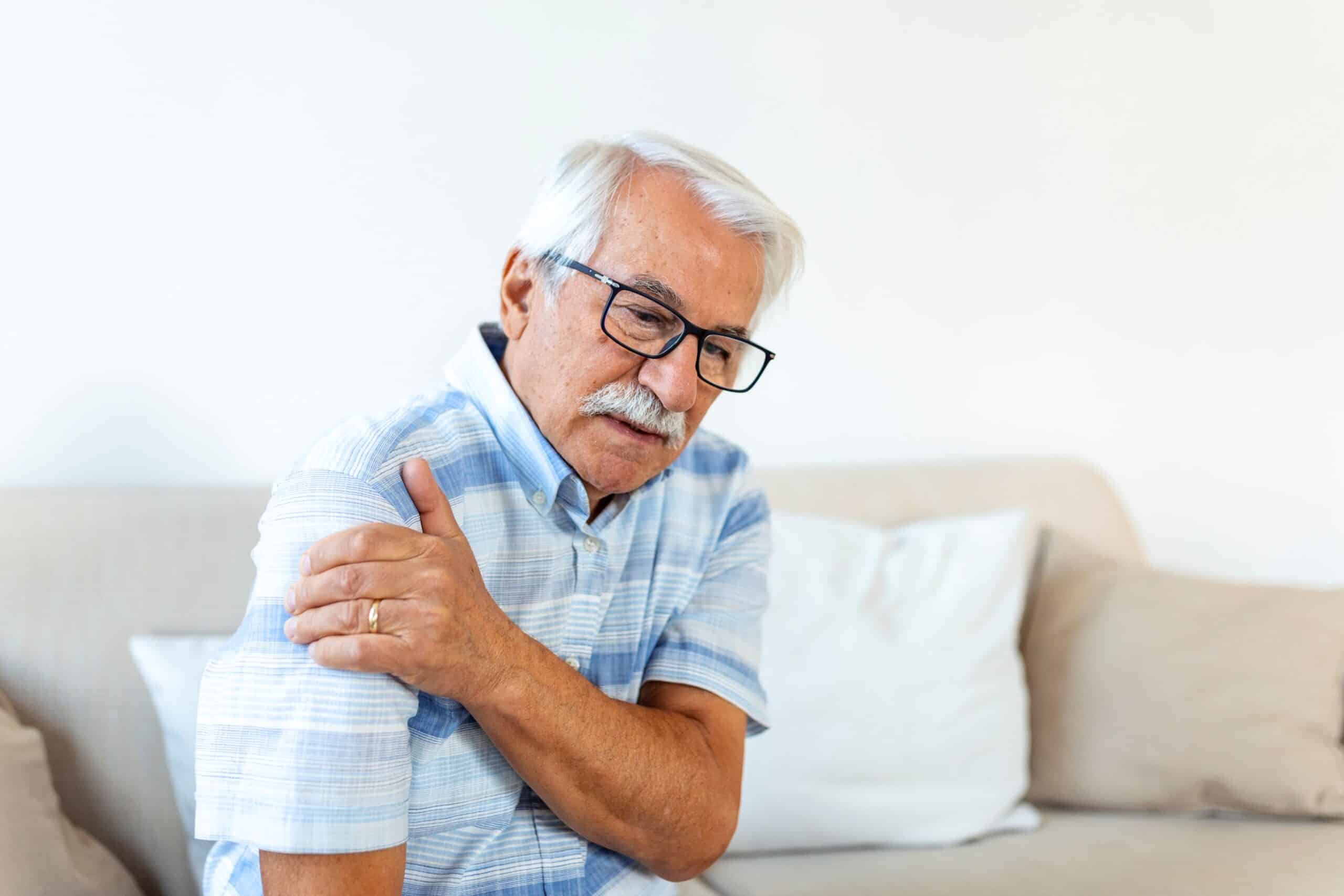 Stages of Inflammation, image of an elderly man sitting on a beige couch, wearing a blue and white stripe button down short sleeve shirt, grasps his right shoulder with his left hand.