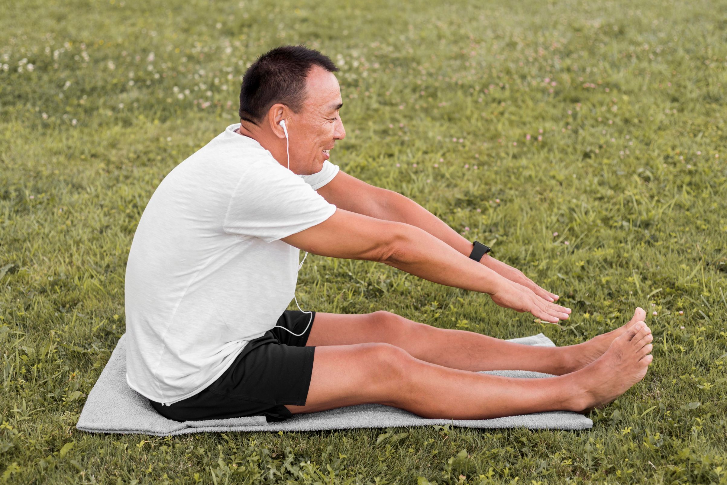 Stages of Inflammation, image of a man wearing a white t-shirt and black shorts sits and stretches his legs on a yoga mat in the grass.